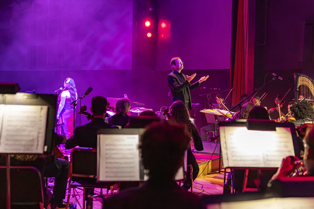A musicians perspective of the WT symphony orchestra performing Hallelujah with a singer soloist. The conductor is leading the musicians against soft purple lighting